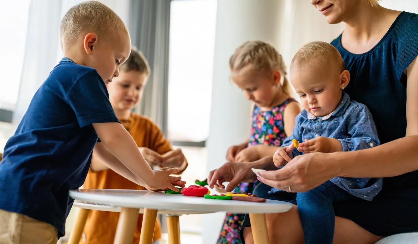 Caregiver guiding infants and toddlers during a sensory play activity at an infant daycare, supporting early development, fine motor skills, and social interaction in a warm, nurturing environment.