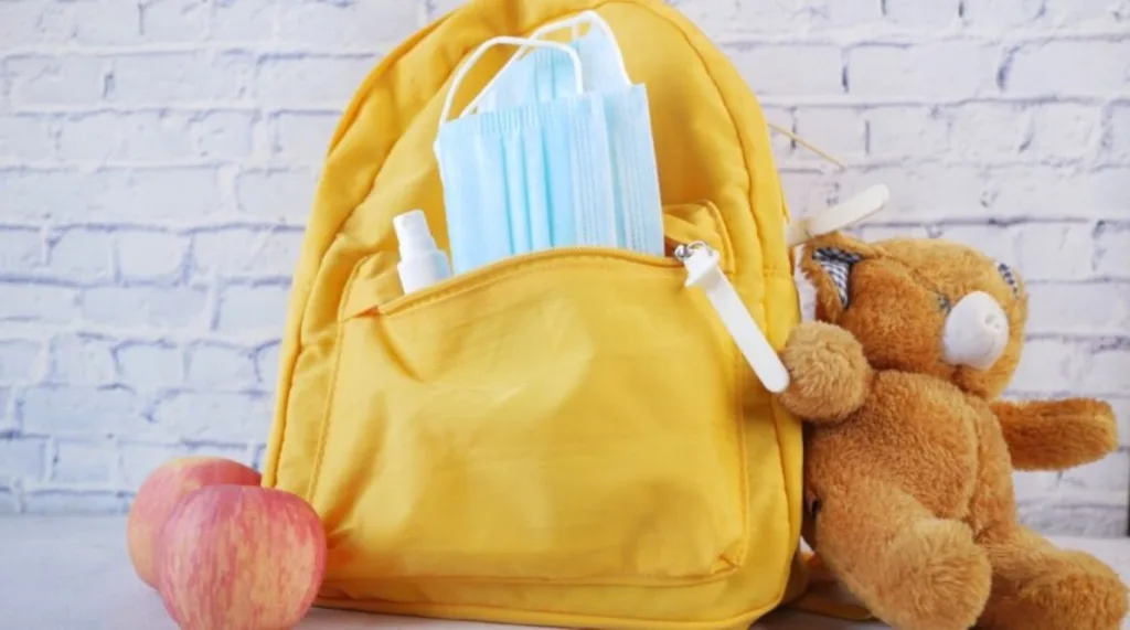 Yellow daycare backpack filled with essential items including a face mask, hand sanitizer, and snacks, placed beside a teddy bear and apple to support a child’s daily routine and comfort at daycare.