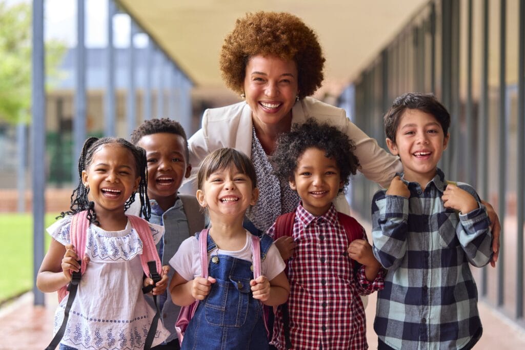 A smiling teacher is embracing a diverse group of toddlers and preschoolers carrying backpacks, showcasing a multiage childcare program with warm, peer learning.
