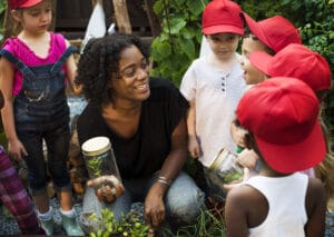 Teacher guiding children during a nature exploration activity at preschools in Langley, where kids observe plants and small insects in jars while learning about the environment outdoors.