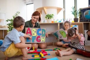 A smiling educator showing a colorful alphabet sign to a group of toddlers during a play-based lesson at a bright and modern early learning center.