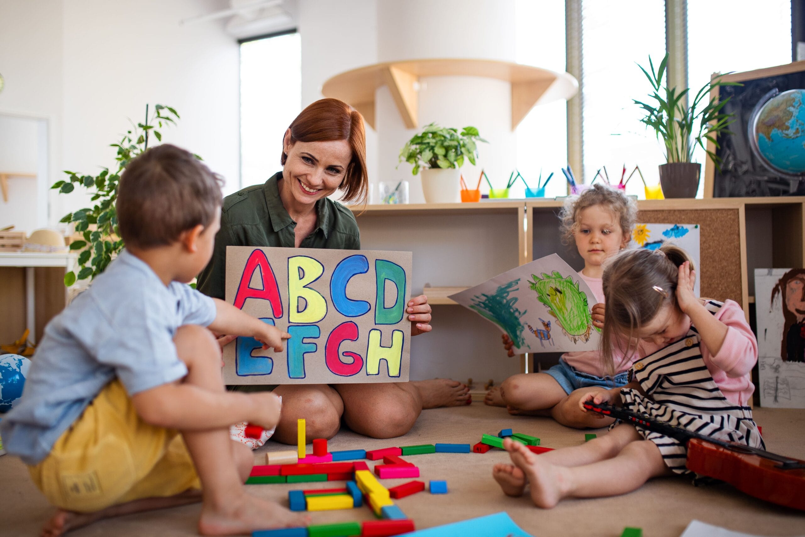 A smiling educator showing a colorful alphabet sign to a group of toddlers during a play-based lesson at a bright and modern early learning center.