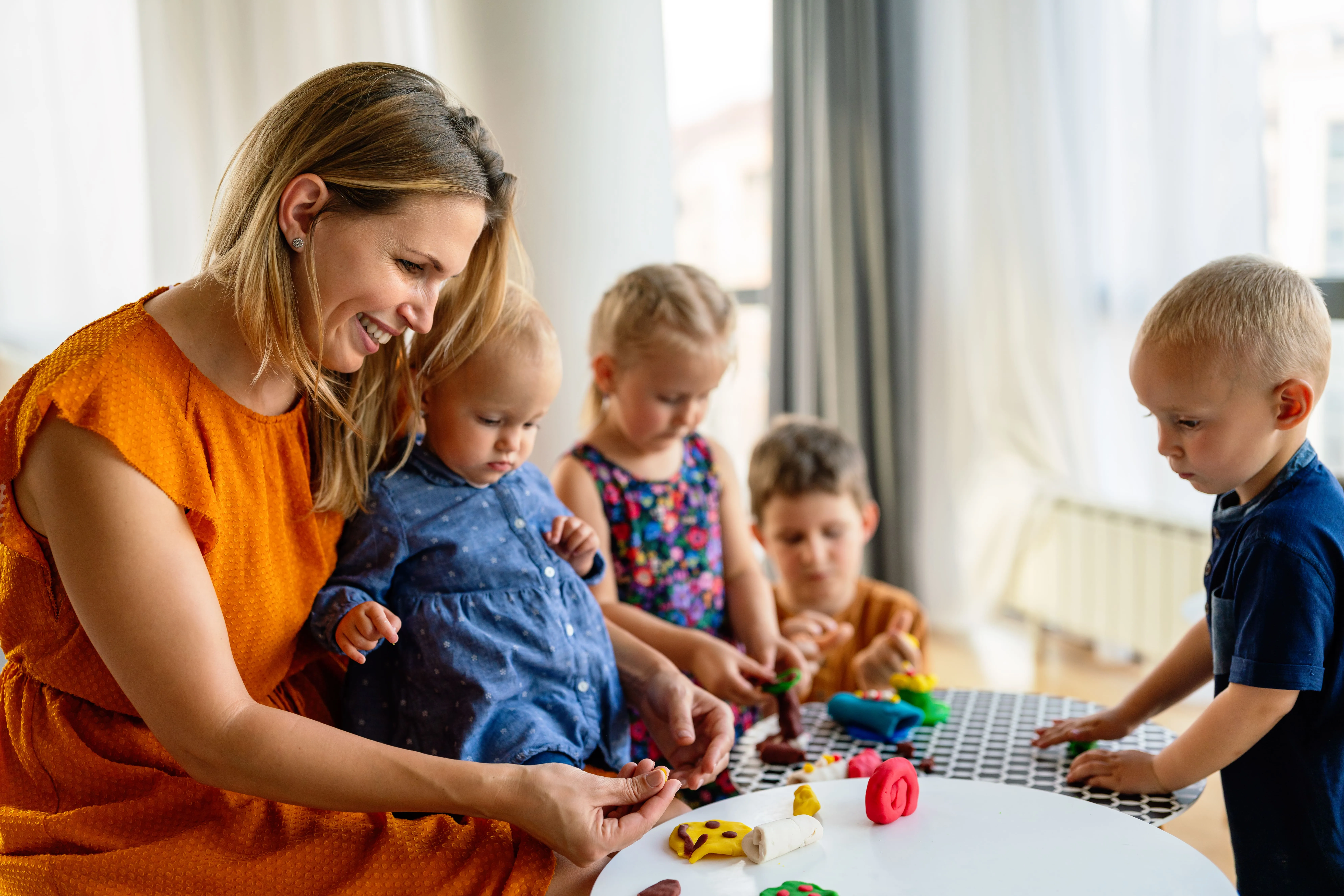 A bright and secure room inside a licensed infant daycare center where a certified early childhood educator is gently supporting a baby during floor tummy time on a clean, colorful foam mat surrounded by soft sensory toys.