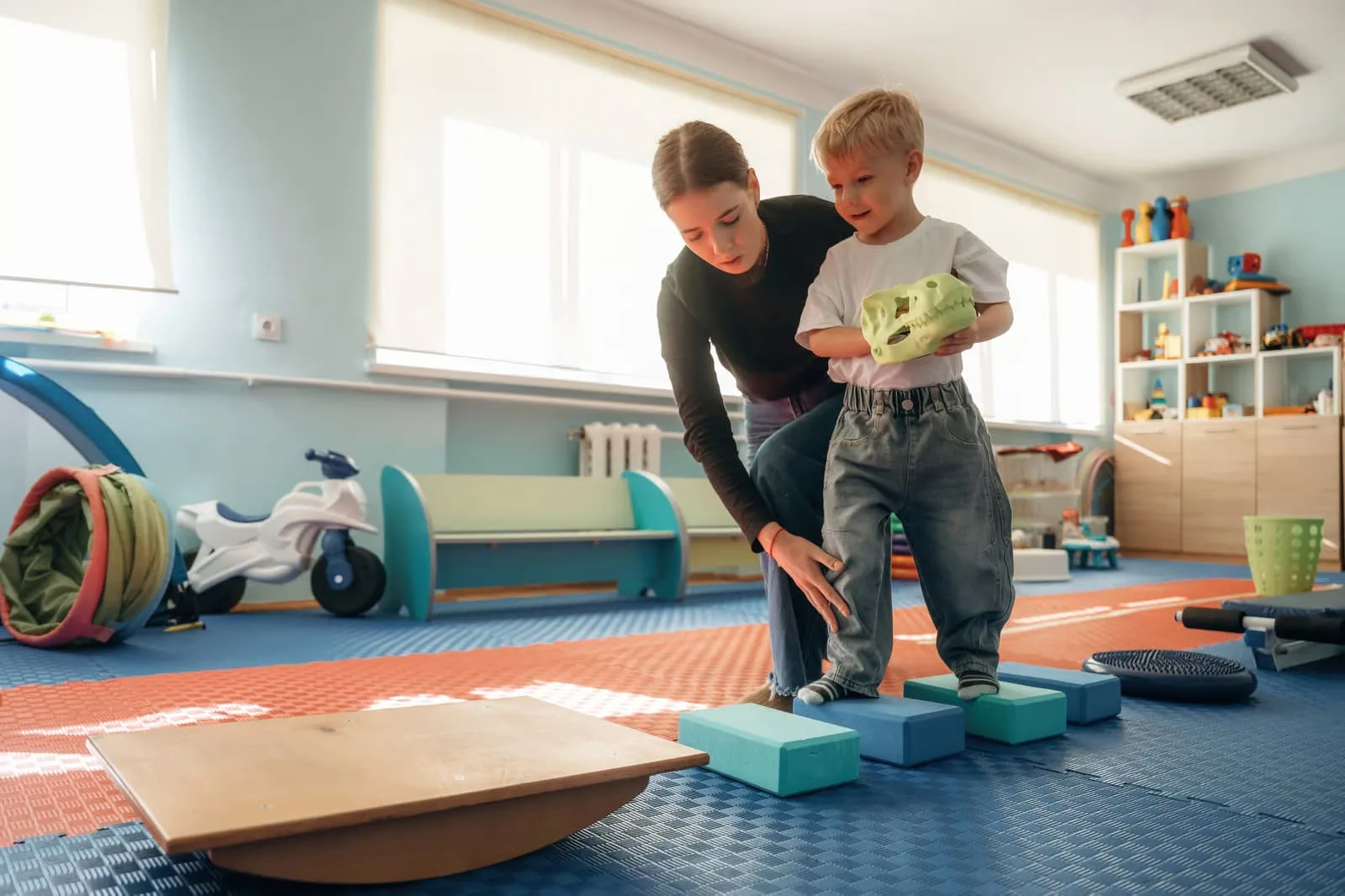 A realistic photograph of a bright daycare center classroom where a young toddler is successfully hanging their backpack on a low wooden hook, demonstrating early childhood independence.