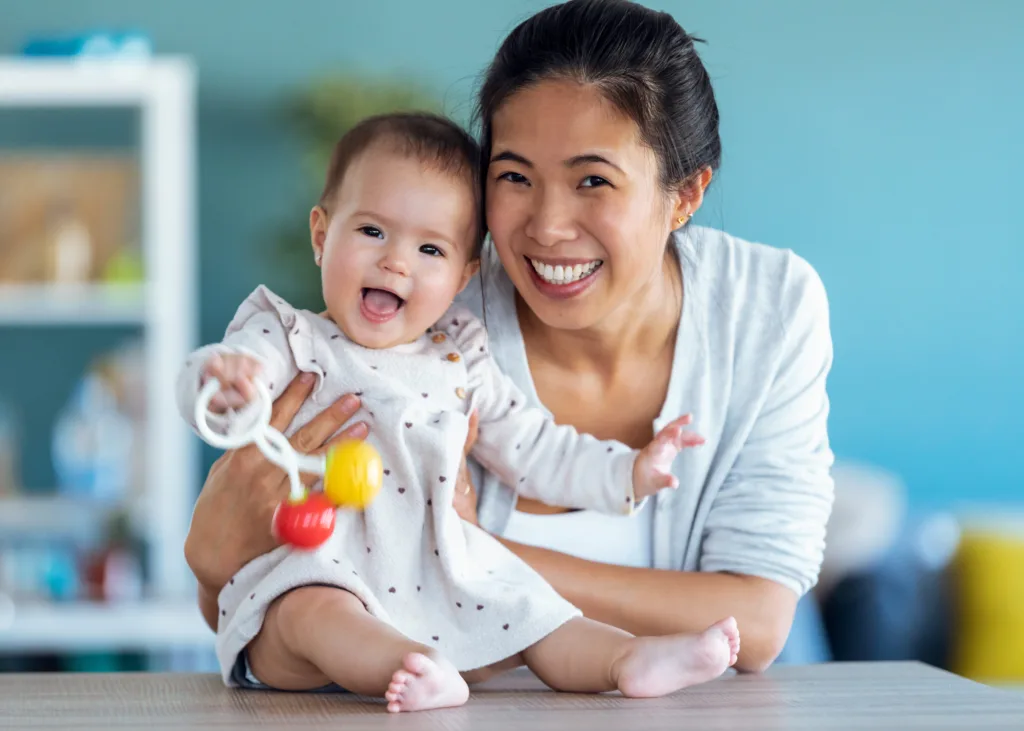 A bright, clean, licensed early learning classroom where a baby is actively exploring a low-level wooden shelf containing natural sensory objects, demonstrating the physical milestone development of infant under the watchful guidance of an educator.