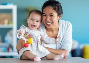 A bright, clean, licensed early learning classroom where a baby is actively exploring a low-level wooden shelf containing natural sensory objects, demonstrating the physical milestone development of infant under the watchful guidance of an educator.